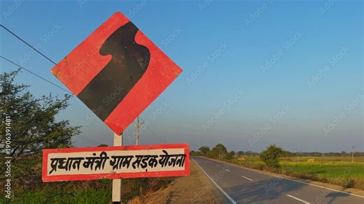 Tracking shot moving past a curve warning sign with Hindi text referencing Pradhan Mantri Gram Sadak Yojana, alongside a rural highway bordered by fields, trees, power lines, and clear blue sky.
