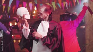 Man disguised as a vampire dances witha skull in his hand. People in different scary costumes are dancing at a halloween party.