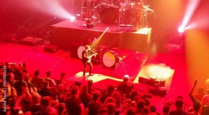 A vibrant concert scene with a large audience enjoying a live performance. The stage is illuminated with red and yellow lights, creating a dynamic and energetic atmosphere.