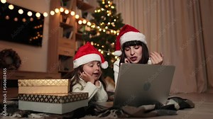 Young caucasian mother and her cute little daughter chatting with somebody using laptop, lying on floor in living room at home decorated with Christmas tree.