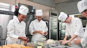 Multiracial professional gourmet team, four chefs in white cook uniforms and aprons knead pastry dough and eggs, prepare bread, and bakery food, baking in oven at stainless steel restaurant kitchen.