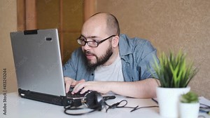 Concentrated adult man geek with beard in eyeglasses typing on laptop, working on project, creating program or trying to crack system, showing yes gesture celebrating victory, freelancer, teleworking