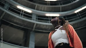 Bottom view of female enthusiast admiring at modern architecture construction of subway. Slow motion. Carefree young black woman look upside on contemporary building with interest