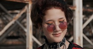 Portrait asian goth girl teenager smiling in sunglasses, leather accessory around neck looking camera, stands against backdrop old abandoned building sun day. Protest. Alternative. Hipster, freak, emo