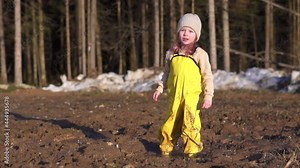 girl have fun in mud. puddle in ground games. Happy child plays in spring village near forest. Yellow waterproof jumpsuit and rubber boots. Splashes of dirt fly to sides and stain clothes. smile baby