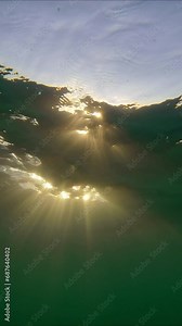 Sunlight underwater at sundown, Verical video, Close-up, Slow motion. Rays of setting sun penetrate under surface of water. Natural underwater background with sunbeams at sunset time