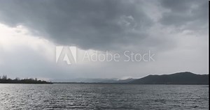 A panoramic view of Mountain Lake Sevan under a cloudy sky. The vastness of the lake is evident as low clouds touch the water, creating a dreamy reflection. Surrounding mountains add to the scene's