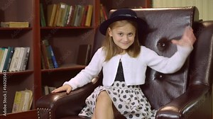 Portrait caucasian little girl five years old sitting on the chair wearing dress and hat posing looking at the camera smiling. Funny baby with long blond hair pose like model.