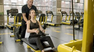 Young woman with instructor working out in gym by using rowing machine