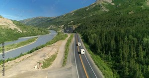 Alaska. Aerial 4K. Camera follows RVs driving along Alaskan mountain road. Calm mountain river is on the left and dense vegetation is on the right of the way. The bends of the road are seen ahead.