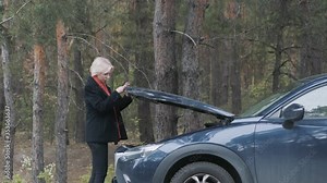 Side view of confident blond woman opening car hood and looking at engine. Adult female driver standing next to broken vehicle next to the forest. Auto accident on countryside road. Lifestyle.