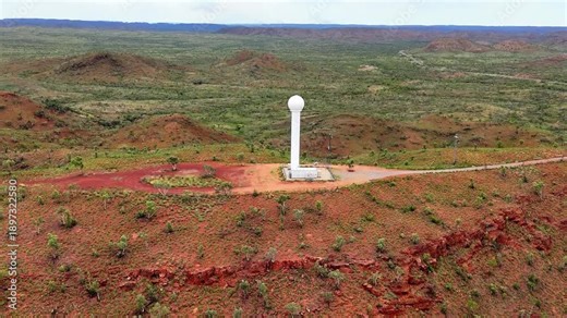 The white weather radar Tower sits beside a small utility complex in arid red-soil landscape. Distant Mount Isa City and sparse vegetation create a contrast between man-made infrastructure and nature