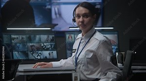 Female operator in headset in police monitoring center turns and looks at camera. Computer monitors and big digital screen with surveillance CCTV cameras video footage on background. Tracking system.
