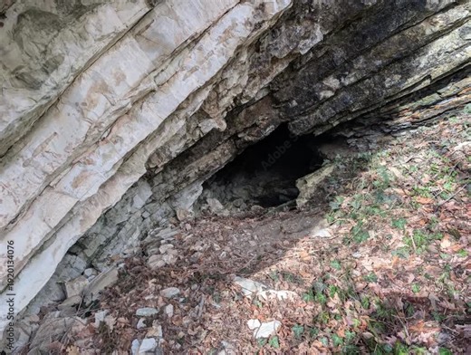 ingresso di una grotta naturale all’interno di una formazione rocciosa stratificata. Le rocce inclinate e fratturate evidenziano processi geologici antichi