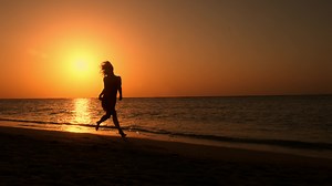 Slow motion woman running barefoot by beach at golden sunset leaving footprints in sand. Female tourist on summer vacation