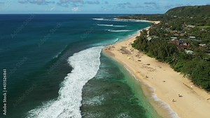 Aerial drone footage above a beach on Oahu, Hawaii, USA. Broad, sandy beach with turquoise water and white waves, and sunbathers visible on the sand. Green forest to the right of the beach.