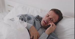Sick young man lying on bed in bedroom at home with seasonal flu or cold sneezing suffering from runny nose and talking by mobile phone checking his temperature feeling unhealthy. 4k video.