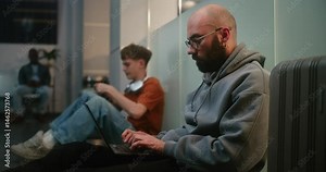 Crowded International Airport Terminal: Man Working Online on Laptop, Sending e-Commerce Emails, Sitting on the Floor in Departure Lounge of Airline Hub. Diverse Passengers Waiting for Delayed Flight.
