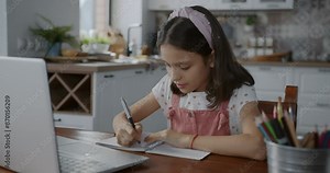 Little girl doing homework writing and looking at laptop screen sitting at table in apartment. Education and childhood concept.