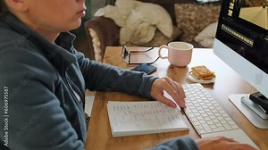 Woman works at home in computer looking at photo on monitor screen and takes notes in notebook. Working at home. Efficiency in modern workplace.