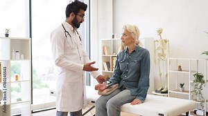 Bearded young male cardiologist telling elderly female patient about all possible reasons of hypertension. Adult woman listening explanations about necessity of leading healthy lifestyle.