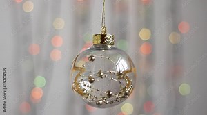 Beautiful Christmas tree toy hangs against the background of multi-colored Christmas lights and moves slightly. Close-up. Transparent Christmas tree toy in the form of a sphere with gold stars and bal