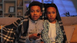 Two little African American brothers are sitting on the sofa covered with a blanket and watching TV. Two multiracial boys get scared while watching a horror movie.