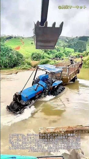 大型ショベルカーが、増水した川で深く泥に埋まったトラクターを救出――泥と水の中での大規模なトラクター引き揚げ作業。チェーンを使って沈み込んだ農用トラクターを引き出すショベルカーの様子。農業機械の泥地か
