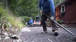 Using a rake upside down, a laborer smooths out the excess gravel that is above the conduit in a hardscaping or landscaping project.
