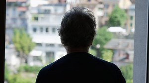 Back of a contemplative senior man looking out window. Pensive older person standing at home staring at view in silhouette