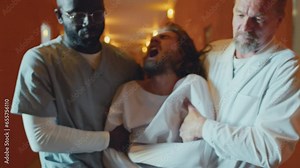 Slow motion shot of two doctors working in asylum restraining violent patient in straitjacket, taking him away along corridor