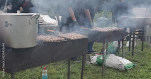 Cooks walking along meat cooking on braziers in a row. Steam and heat around. Men in black uniform walking around while cooking for summer party outdoors. Braziers with shish kebab on coal.