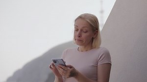 A crying woman wiping her tears uses a smartphone on the balcony of her hotel room overlooking the sea.