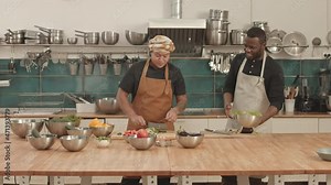Medium long of young African American female and male sous-chefs wearing aprons, standing at kitchen island, cutting fresh cucumber and eggplant on cutting boards, talking and smiling