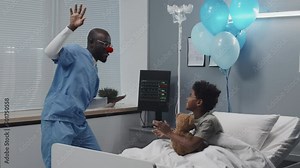 Medium long of young male nurse in clown nose and scrub suit making faces and entertaining ill African American boy who sitting in hospital bed with stuffed bear, smiling and clapping hands