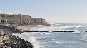 View of the coast from the city of Cádiz in Andalusia, Spain. Waves hit the coast. People are walking on the city's beaches in front of the buildings. In the foreground there are rocks. Time lapse.