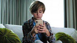 Handsome caucasian boy kid using mobile phone, chatting. Teenager holding phone gadget for playing games, chatting, sitting alone on the sofa in living room