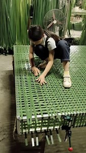 They are making the mat by weaving green plastic strips together in a grid pattern. The girl is crouching on the mat, using her hands to place and arrange the strips. The mat is built on a metal frame to keep it in shape. Rolls of green strips and a fan can be seen in the background, showing that the work is done by hand in a warm workshop. | Nature View