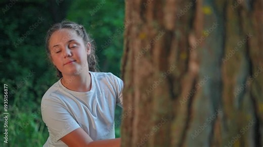 Teenage girl stands near tree trunk in forest park outdoors. Young woman crosses arms beside tree in nature. Teen girl smiles near tree. Portrait of teenager in park. Girl stands by forest tree.