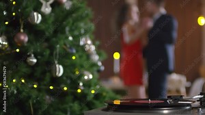 Close-up shot of vinyl record playing on turntable, Christmas tree with decorations and lights, and man and woman in red dress dancing together and swaying to music in blurred background