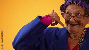 Funny closeup view of smiling happy elderly senior old woman with wrinkled skin smiling pointing and giving call me gesture with hands isolated on solid yellow background.