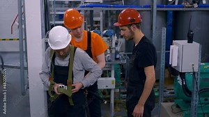 A bearded male engineer shows workers how to put on a safety belt, heavy industry workers and workers in uniform stand at a petrochemical plant. Industrial contractor trains staff