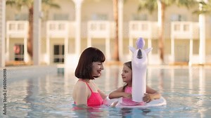Little girl with her mom having fun in swimming pool. A young mother teaching her daughter swim in clear blue water in a pool with an inflatable lifebuoy in the form of a unicorn.