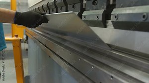 A big machine is bending a long thin metal item in the factory. A worker is holding it with safety gloves. Close-up shot.