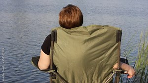 Fisherwoman catching fish. A woman is sitting on a folding tourist chair on a riverbank with a fishing rod in hands, catching fish. Wooden pier, green reeds on a bank. Water flowing. Leisure weekend.