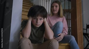 Dissatisfied Caucasian boy closing ears as displeased girl yelling sitting on stairs at background. Portrait of sad brother looking at camera as furious sister scolding sibling indoors at home