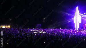 People in the stadium in front of the stage. Spotlights illuminate people in the stadium. Thousands of people in the stadium listen to music and dance. Rays of light from the stage. Many people dance