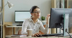 Young tired woman in glasses sitting at the laptop computer while working in the office, then almost falling asleep and waking up. Indoor.