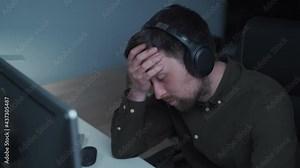 Young caucasian man working overtime at computer and eye fatigue from monitor in home office. Male having stressful time working on PC. Exhausted programmer stressful work, stress at workplace