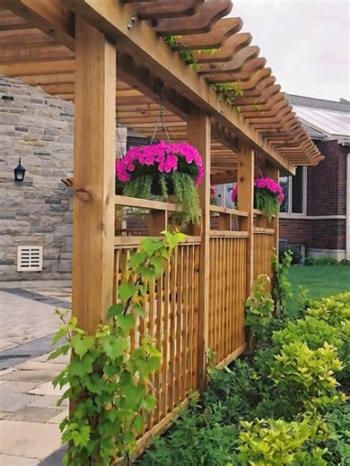 a beautifully crafted, custom wooden pergola-style privacy screen that serves as a functional and aesthetic transition between a paved patio and a lush garden area. STRUCTURAL DESIGN The structure is built from high-quality, warm-toned wood (likely cedar or redwood) and consists of three main sections: • Vertical Support: Robust square posts provide the primary framework, anchored firmly into the ground along the edge of the patio. • Privacy Panels: The lower two-thirds of the structure feature 